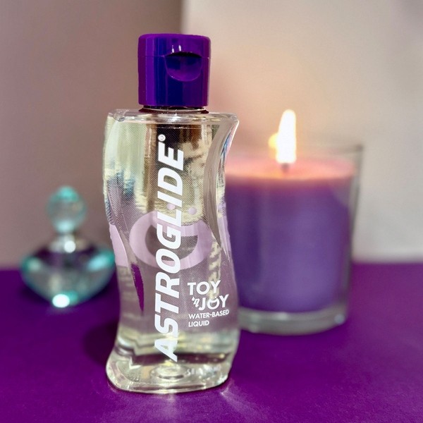 A bottle of Astroglide Toy 'N Joy Lubricant with a purple cap sitting on a purple surface, next to a lit purple candle.