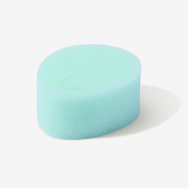 A studio photograph of a single light mint green sponge tampon against a white background. The sponge has an oval shape and a porous texture, with a subtle crescent-shaped indentation on the top surface used for removal.