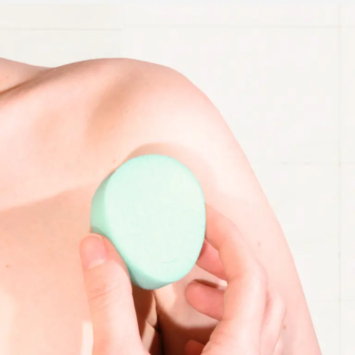 A lifestyle photograph showing a close-up of a person's hand holding a mint green sponge tampon against their upper thigh. The lighting is bright and clean, emphasizing the soft texture of the sponge against the skin.