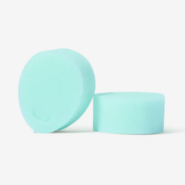 A studio shot of two mint green sponge tampons. One sponge is positioned upright on its side to show its height and the removal loop indentation, while the other sits flat next to it. The background is a minimalist, solid white.