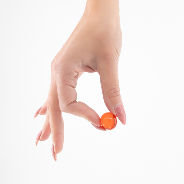 Close-up of a hand with manicured pink nails delicate holding one small translucent orange oil ball between the index finger and thumb against a white background.