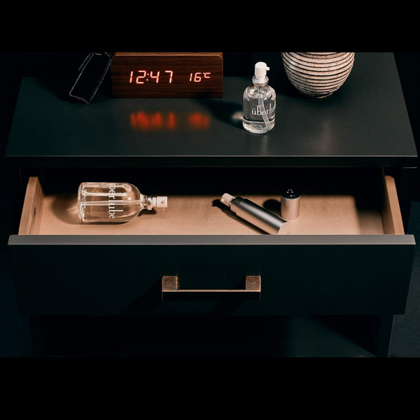 
An open nightstand drawer reveals an Überlube bottle and a silver applicator, while another Überlube bottle, a digital clock displaying "12:47 16°C," and a textured vase rest on top. This discreet arrangement showcases practical and intimate products available through WorldCondoms, ready for everyday convenience.