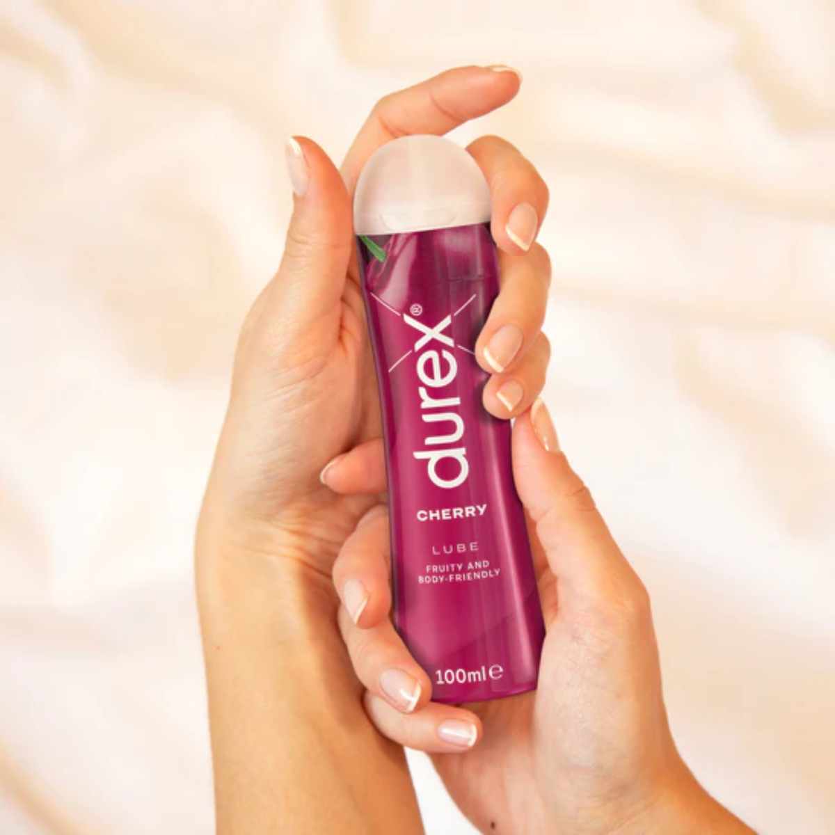 A pair of hands holding a pink 100ml Durex Cherry Lube bottle against a background of white sheets. The bottle is labeled "CHERRY LUBE FRUITY AND BODY-FRIENDLY."