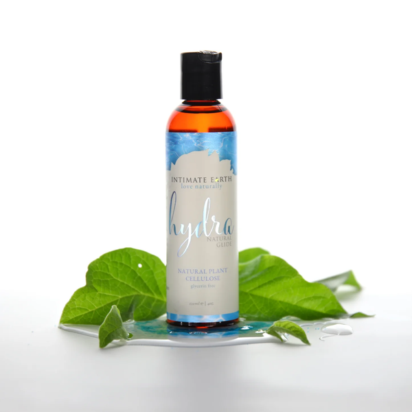Nature-inspired studio photo showing the 120 ml bottle of Intimate Earth Hydra natural plant cellulose glide resting on a wet reflective surface surrounded by green leaves.