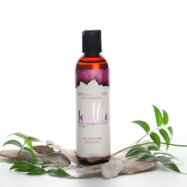 Nature-inspired lifestyle photo showing the Intimate Earth Soothe 120 ml amber bottle resting among natural tree bark pieces and fresh green leaves.