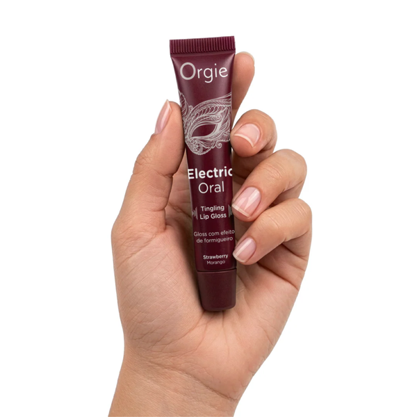 A close-up shot of a hand with neat, naturally manicured fingernails holding the small, maroon Orgie "Electric Oral" lip gloss tube to provide a sense of scale. The tube is held vertically between the thumb and fingers against a plain white background.