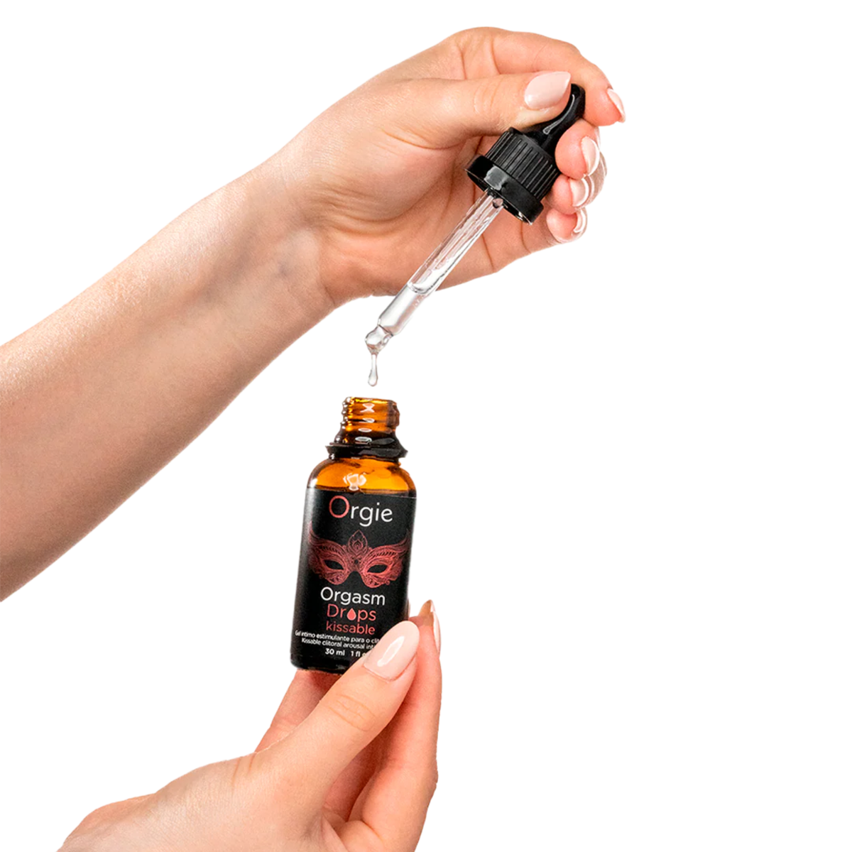 A person's hands holding an open 30ml amber glass bottle of Orgie Orgasm Drops against a white background. The right hand holds a glass dropper, showing a clear liquid droplet suspended from the tip, while the left hand stabilizes the bottle. The black label features a red masquerade mask design and the text "Orgasm Drops kissable".