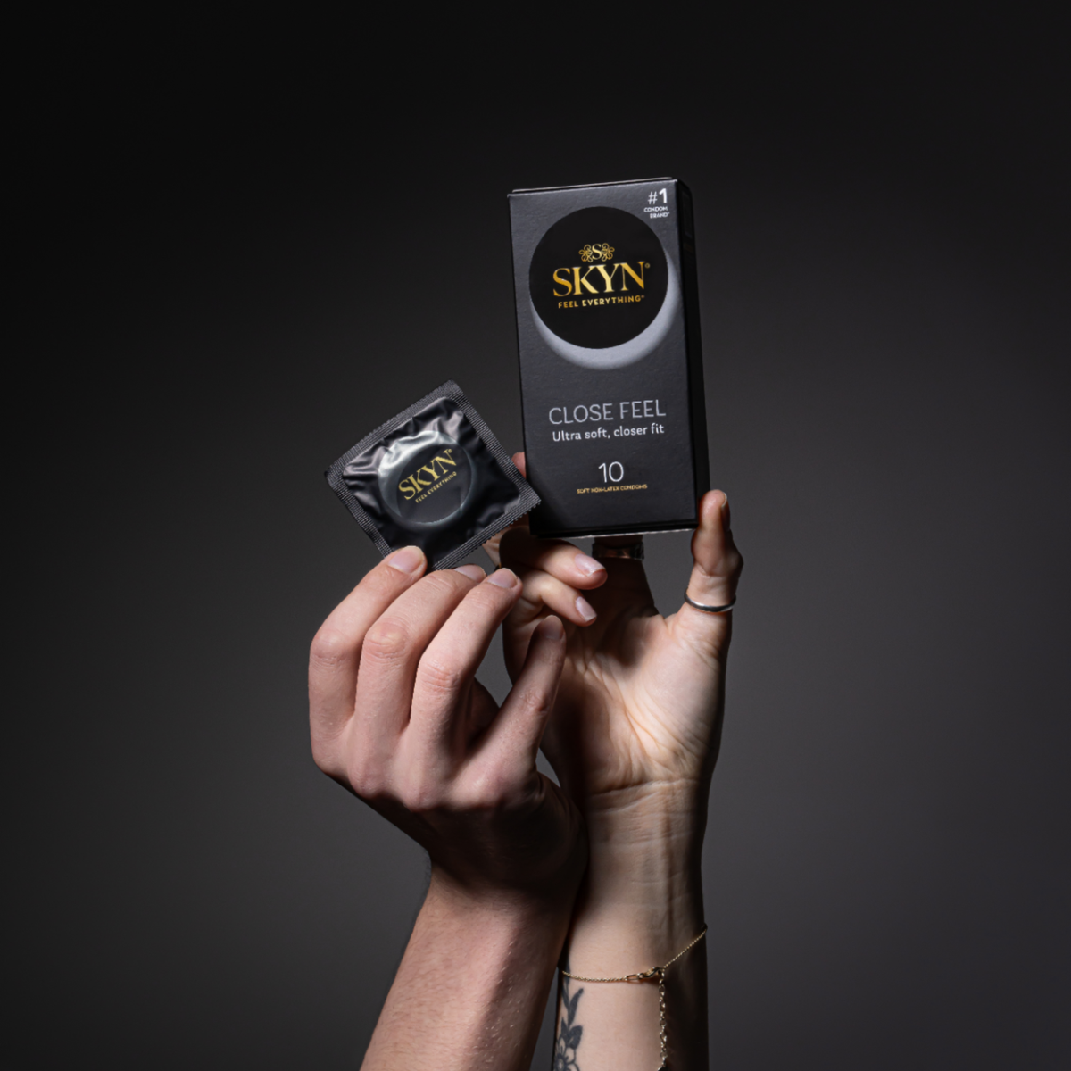 Two hands reaching upward against a dark grey background, holding a black box of SKYN Close Feel condoms and a single black foil condom wrapper. The box indicates it contains 10 soft non-latex condoms and features the "Feel Everything" slogan. Both the box and the wrapper prominently display the gold SKYN logo.