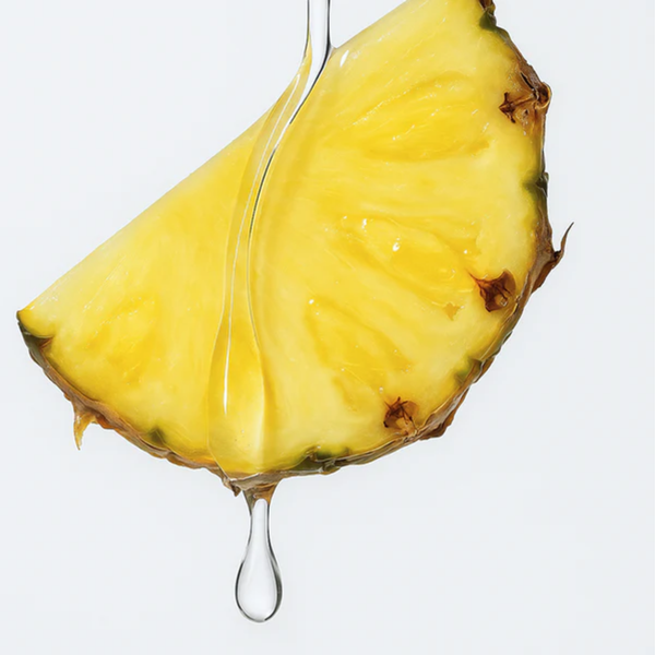 A macro close-up shot of a single juicy slice of yellow pineapple against a clean white background. A thick, clear drop of lubricant slowly trickles down the front edge of the fruit.
