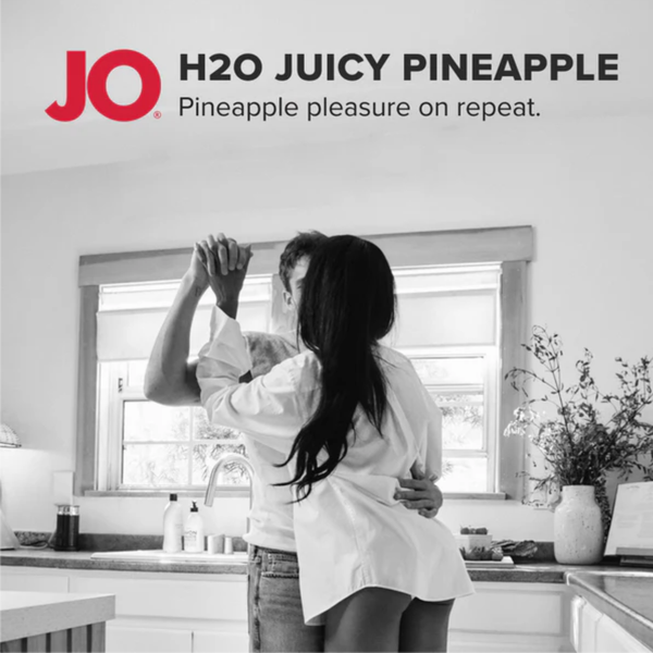 A black and white photograph of a happy couple slow-dancing together in a bright kitchen. The red "JO" logo stands next to black text introducing the flavored lubricant as "Pineapple pleasure on repeat."
