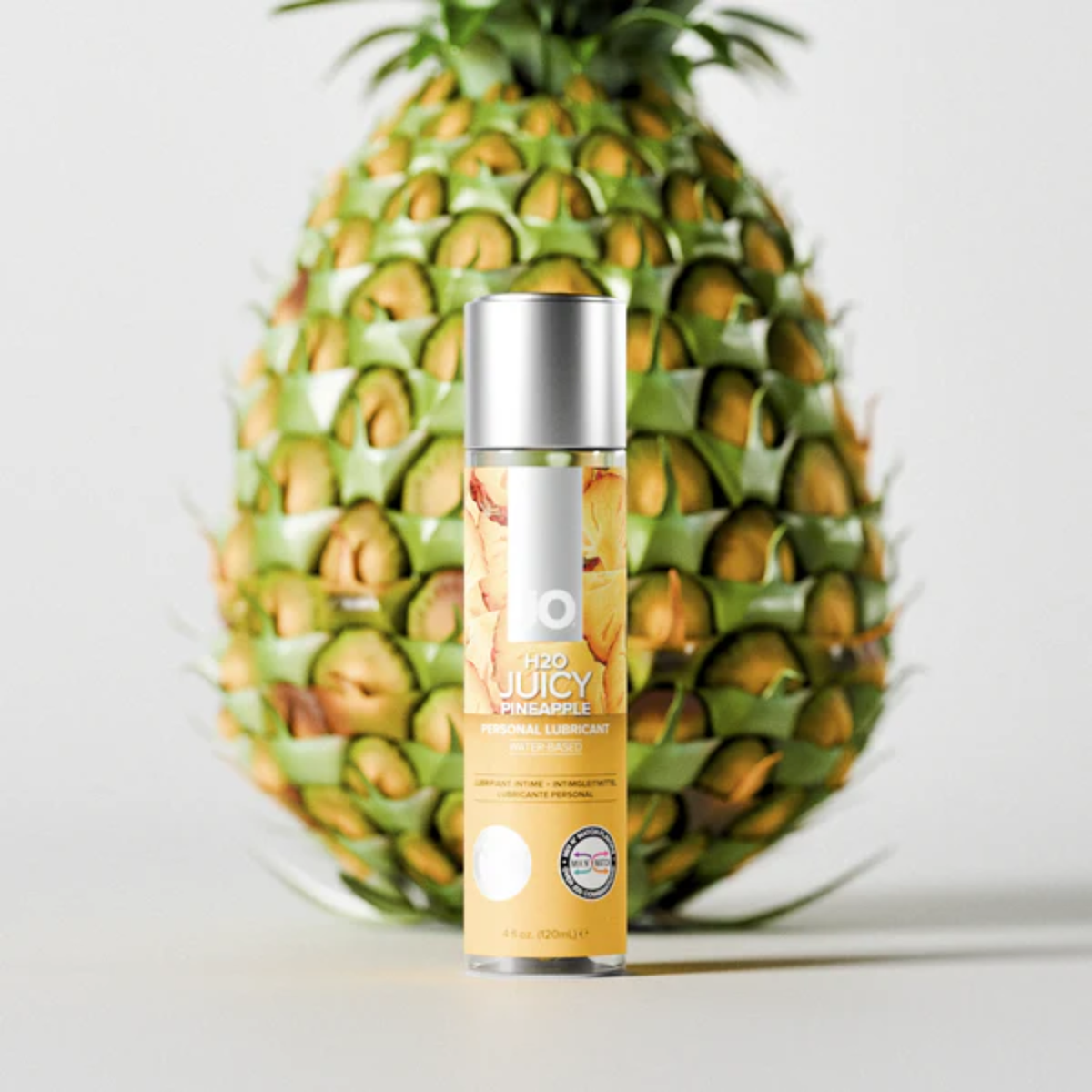 A bottle of System JO H2O Juicy Pineapple lubricant positioned vertically in front of a whole, large pineapple. The entire arrangement stands centered against a smooth white studio backdrop.