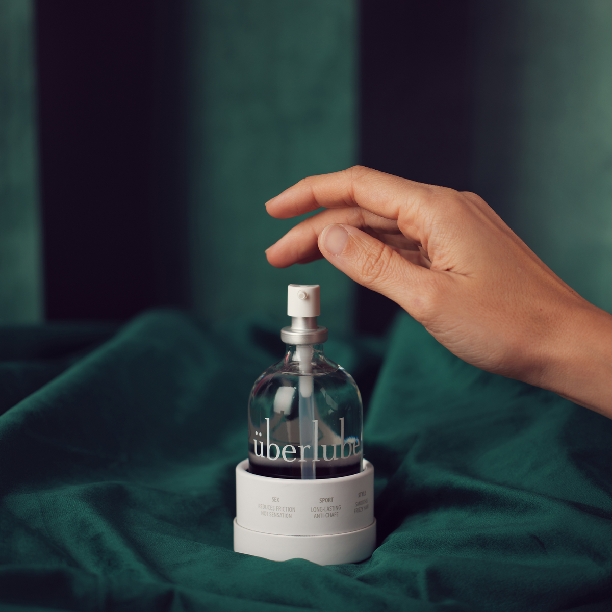 A hand reaches for a clear glass bottle of Überlube Luxury Lubricant on a white base, set against a dark green velvet background. The bottle has a silver pump top and the word "überlube" is printed in white on the lower part of the bottle. From worldcondoms assortment.