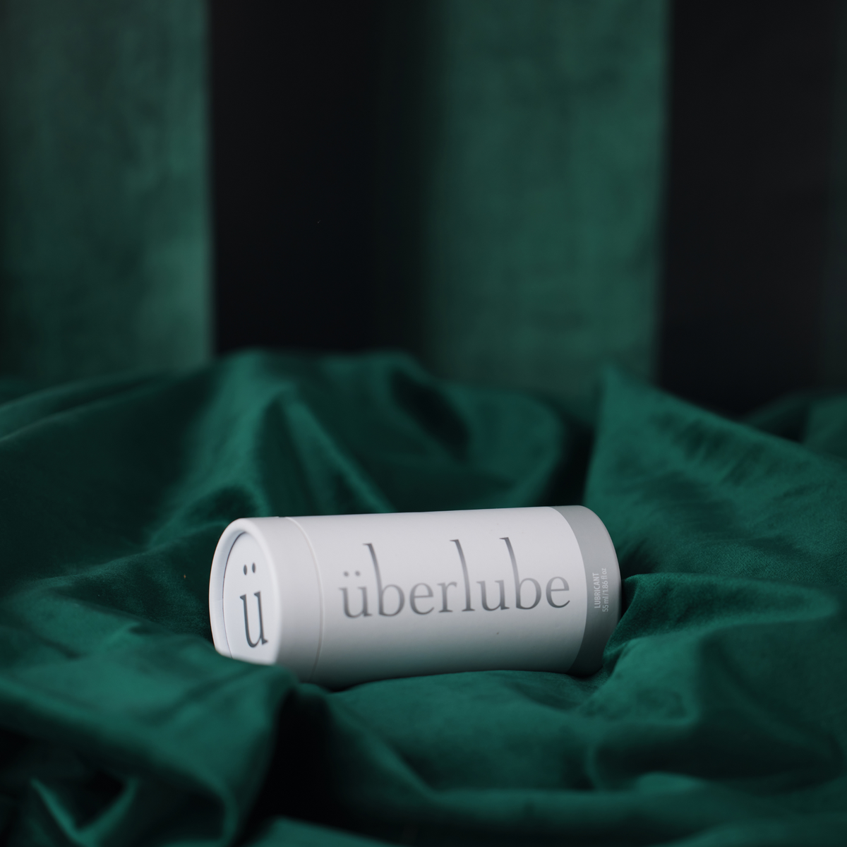 A horizontal shot shows a white tube of Überlube lubricant, with its cap tightly closed, lying on a swath of dark green satin or velvet fabric. From worldcondoms assortment.
