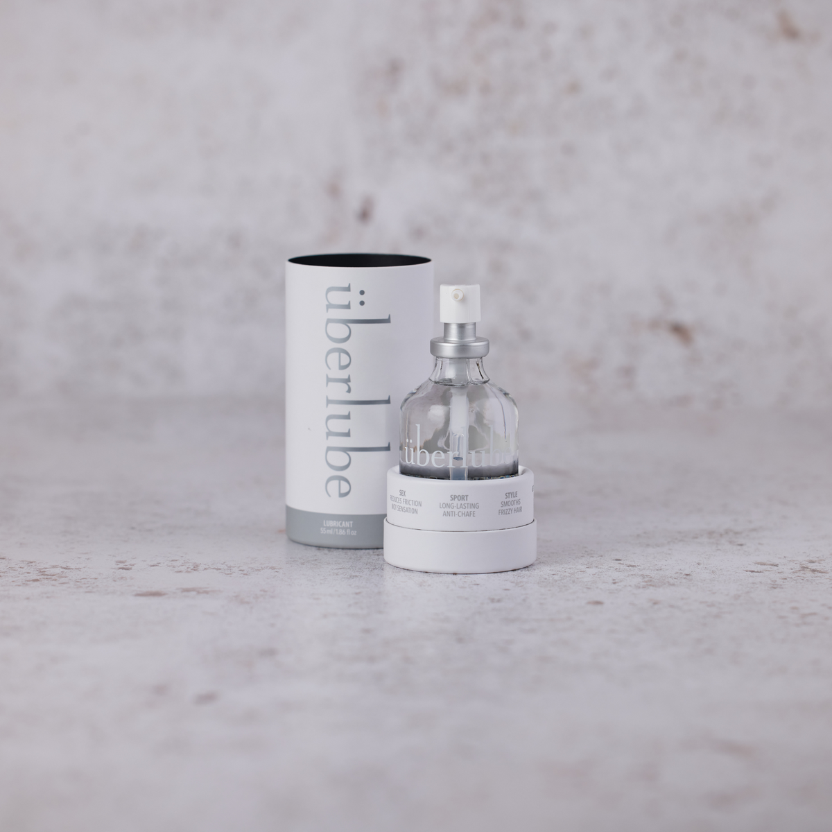 A clear glass bottle of Überlube lubricant with a white base and silver spray pump stands in front of its cylindrical white packaging tube, displaying the "überlube" logo. This clean and modern presentation reflects the quality products available from WorldCondoms.




