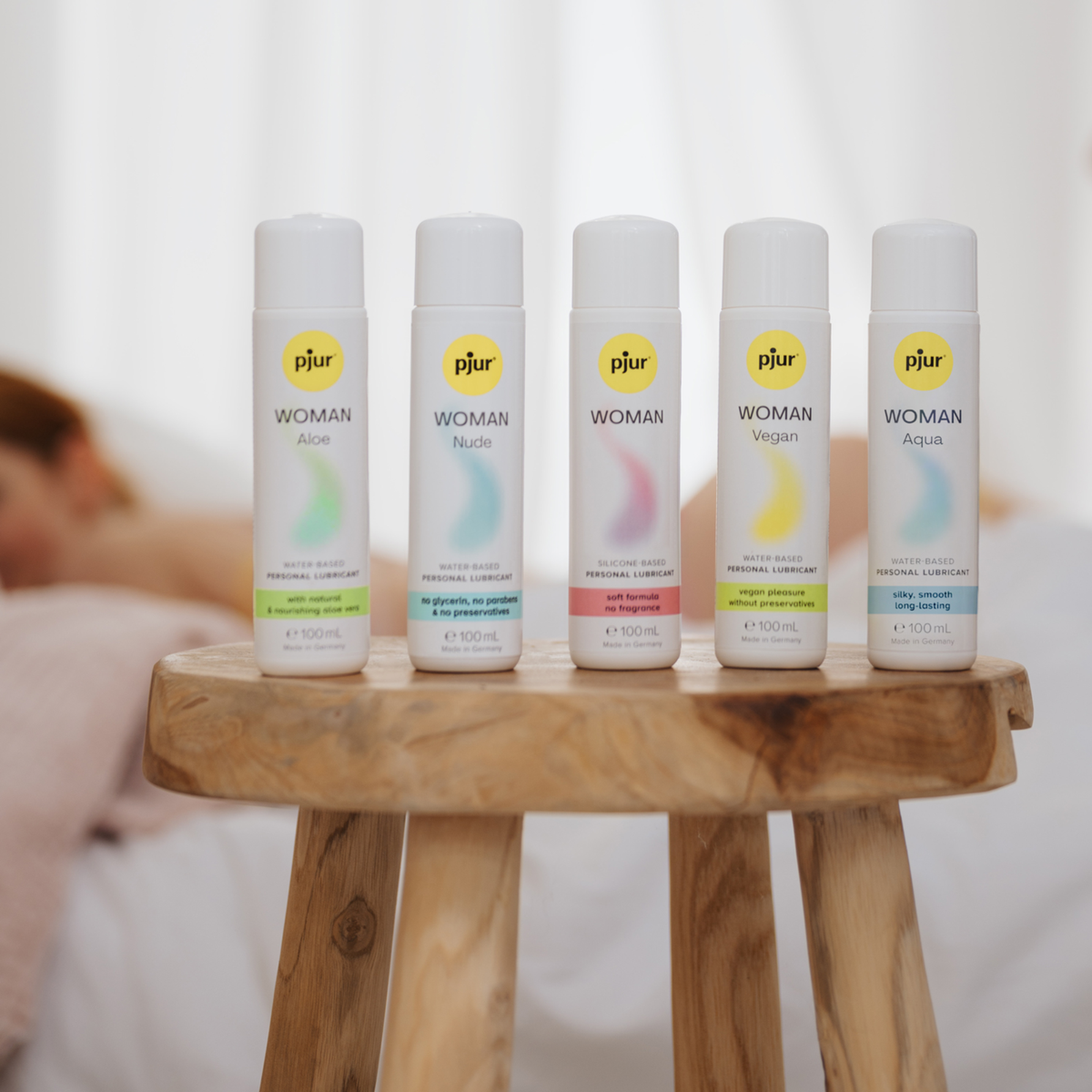 A product photo showing five bottles of pjur WOMAN personal lubricant lined up on a wooden stool. From left to right, the variants are: Aloe, Nude, Woman, Vegan and Aqua.