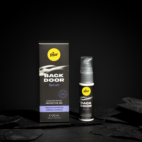 A gray box and a clear pump bottle of Pjur Back Door Serum are positioned against a dark background with textured dark gray elements. 