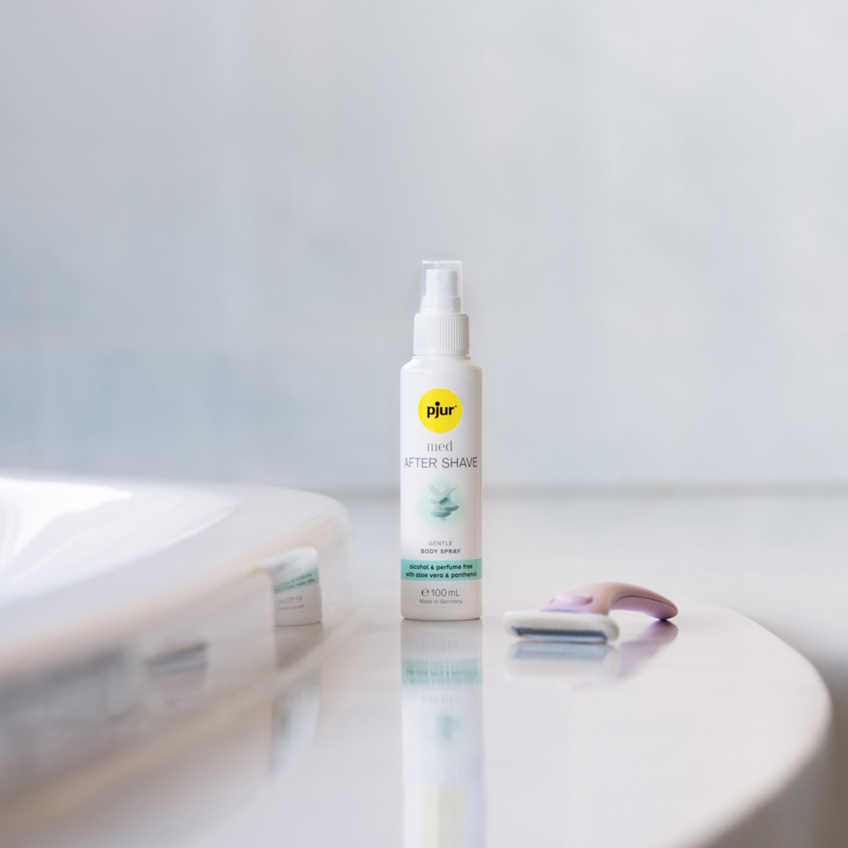 A white bottle of pjur med AFTER SHAVE spray stands on a white surface next to a pink and grey razor, with a blurred white object in the foreground and a light grey background.