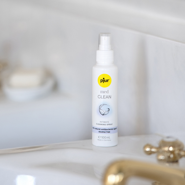 A white spray bottle of pjur med CLEAN intimate cleaning spray stands on a white surface near a blurred white sink and gold-toned faucet.