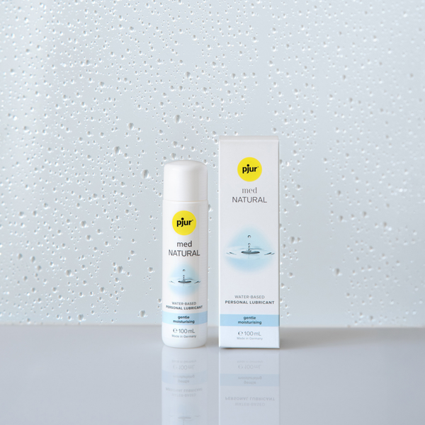 A white bottle and box of pjur med NATURAL water-based personal lubricant stand on a white surface against a background with water droplet patterns.