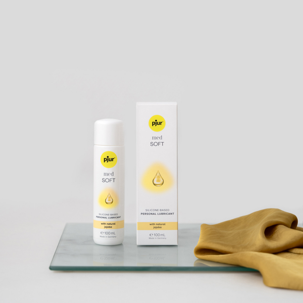 A white bottle and box of pjur med SOFT silicone-based personal lubricant are positioned on a clear glass surface with a piece of soft, golden fabric. The background is plain white.