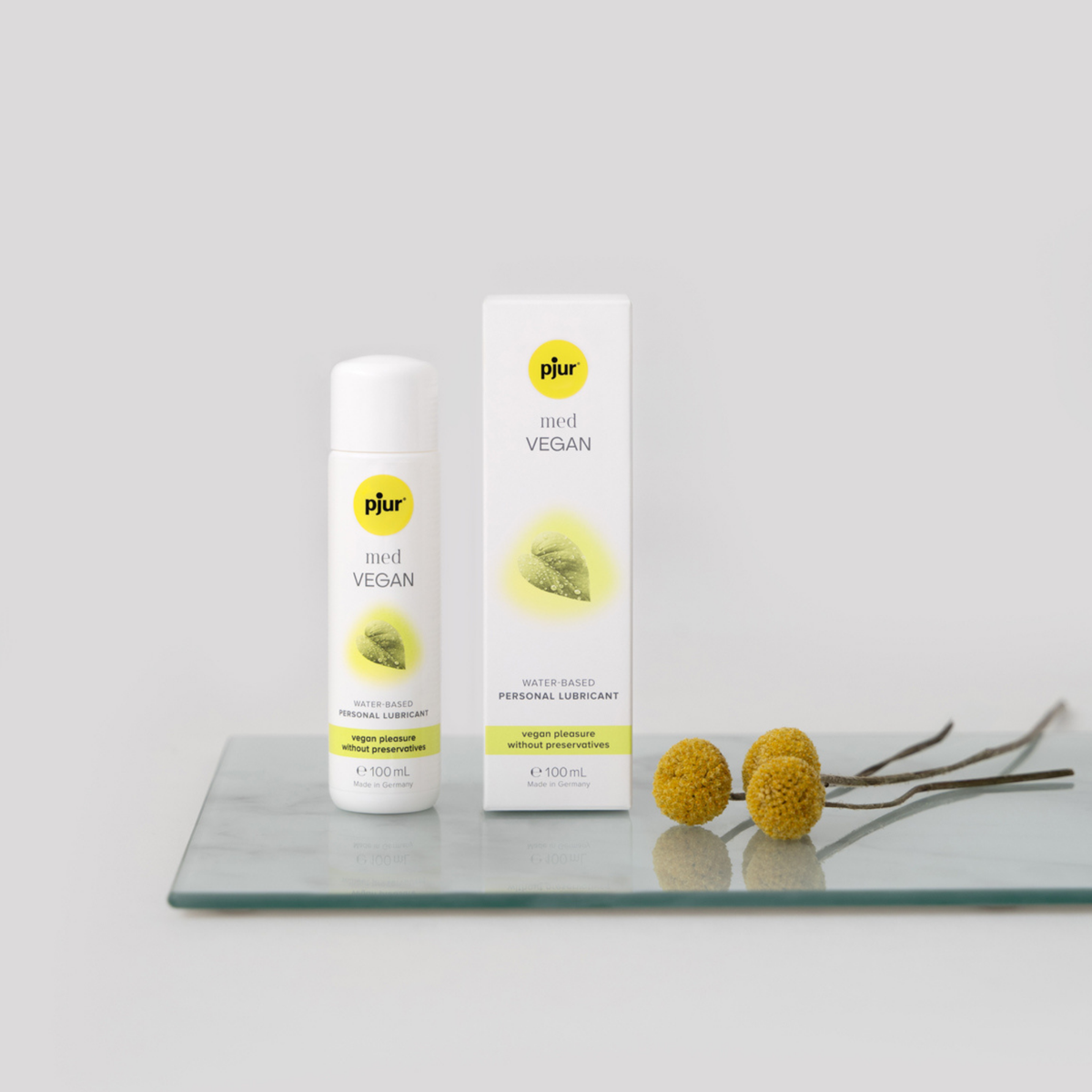 A white bottle and box of pjur med VEGAN water-based personal lubricant are displayed on a clear glass surface with two small yellow flowers and their stems. The background is a soft grey.