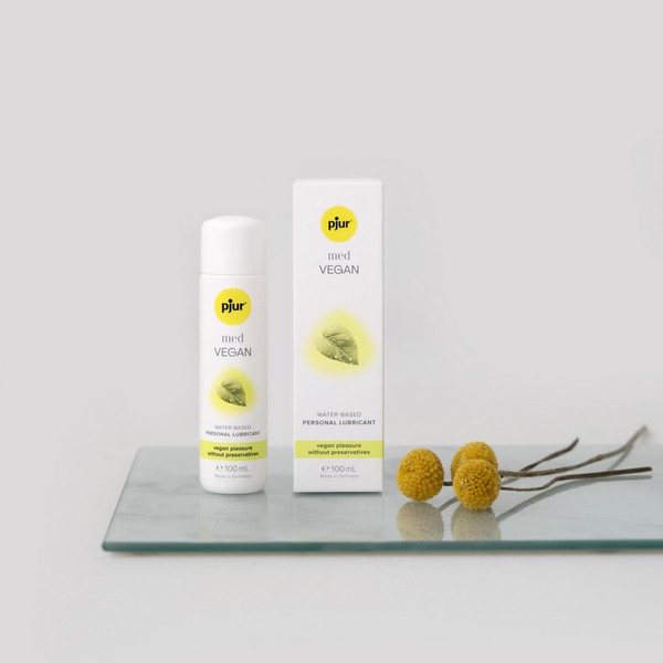A white bottle and box of pjur med VEGAN water-based personal lubricant are displayed on a clear glass surface with two small yellow flowers and their stems. The background is a soft grey.