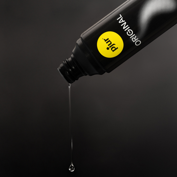 A black bottle of Pjur Original lubricant is tilted, with a clear drop falling from its nozzle against a dark background.