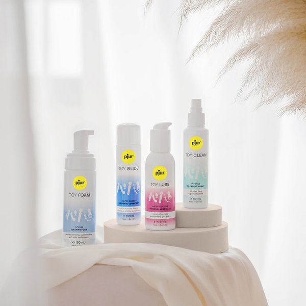 Four white bottles of Pjur toy care products are arranged on small white platforms draped with a sheer white fabric.