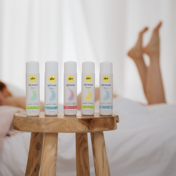 Five white bottles of Pjur Woman products variants are arranged on a wooden stool near a bed with partially visible legs.