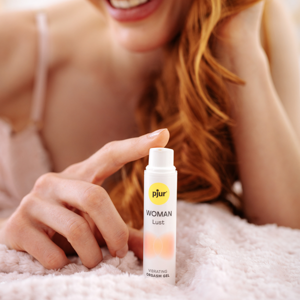 A person with fair skin and reddish-blonde hair holds a tube of pjur WOMAN Lust vibrating orgasm gel on a soft, textured surface.