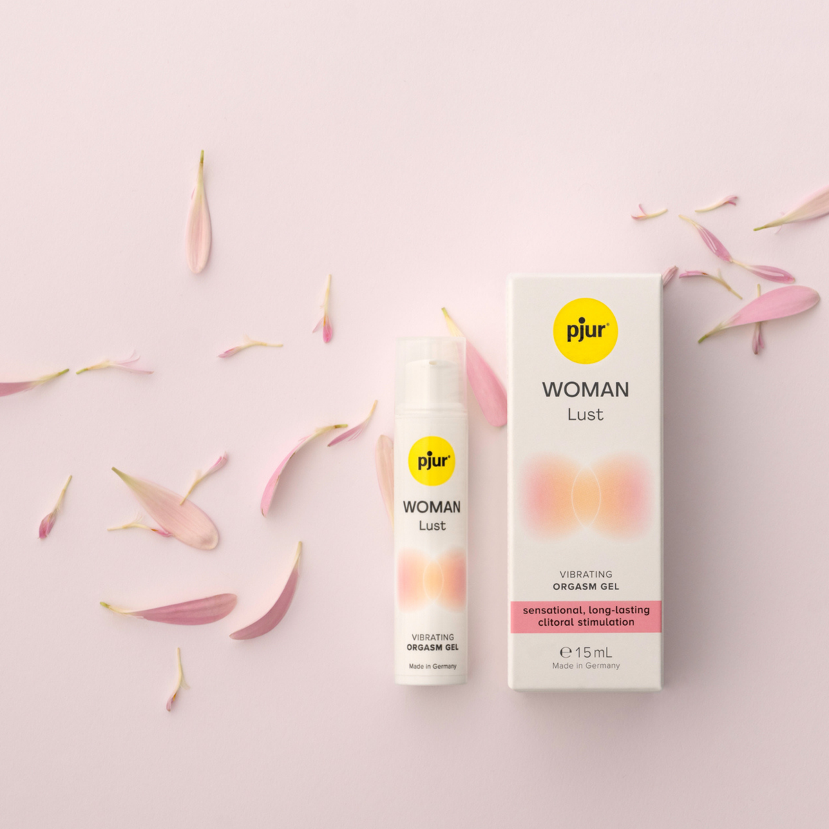 A tube and box of pjur WOMAN Lust vibrating orgasm gel surrounded by scattered pink flower petals on a light pink background.