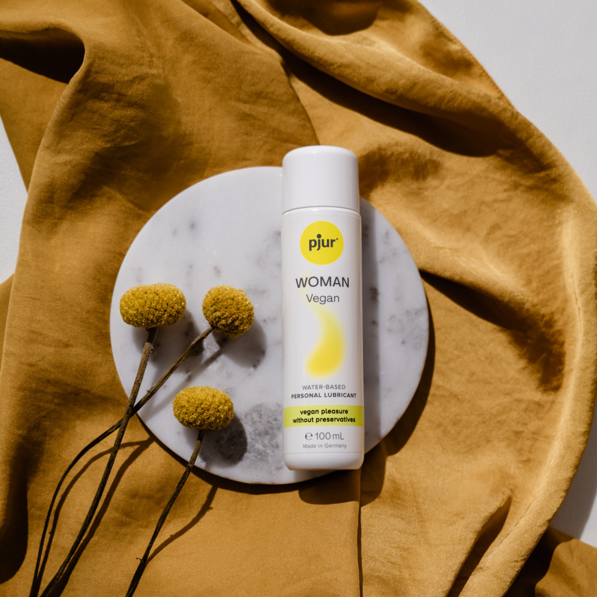 A white bottle of Pjur Woman Vegan lubricant rests on a marble coaster with dried yellow flowers, against a draped mustard-yellow fabric.