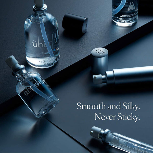 This close-up shot features multiple clear glass Überlube lubricant bottles, some with their caps off, arranged on a dark, sleek surface. The text "Smooth and Silky. Never Sticky." is prominent, highlighting a key benefit of this WorldCondoms lubricant.