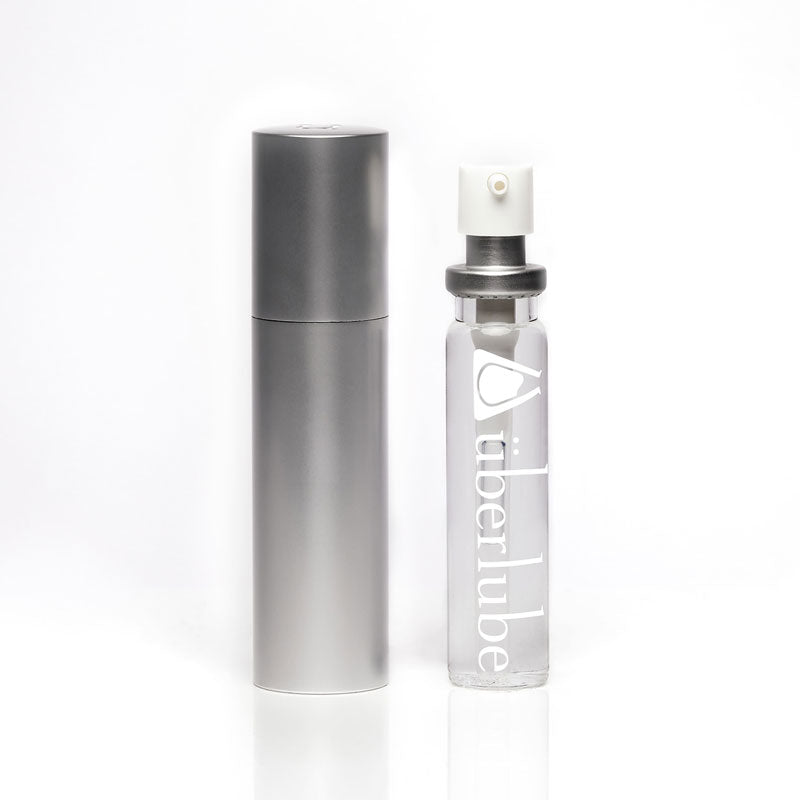 A tall, slender silver refillable case stands next to a clear glass bottle of "überlube" with a white pump top, both items for personal lubricant available at Worldcondoms.