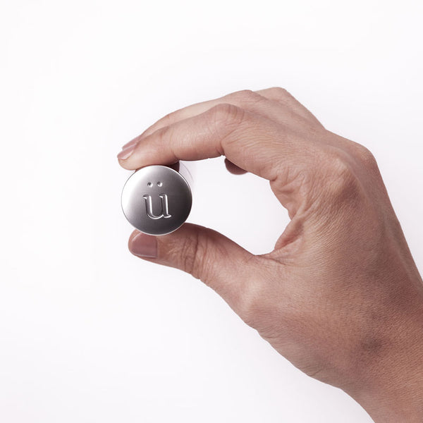 A hand holds a sleek, silver, cylindrical dispenser with a white pump top, likely a personal lubricant from Worldcondoms.