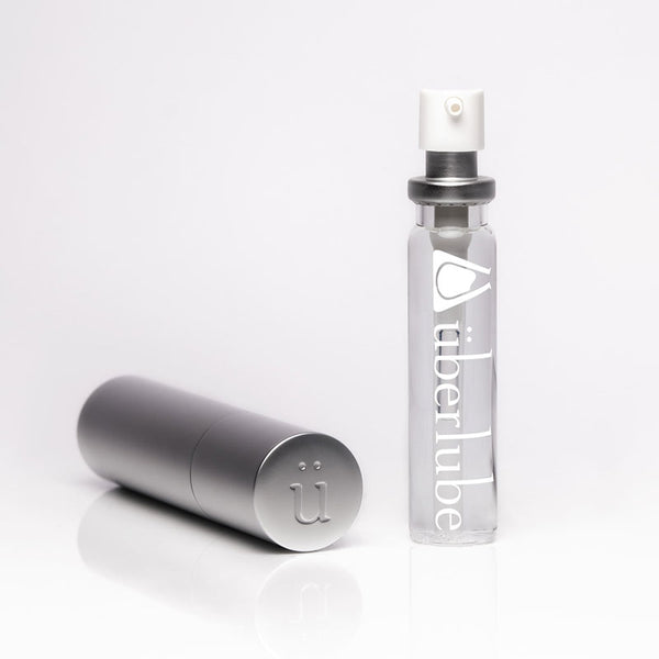 A clear "überlube" bottle with a white pump top stands upright next to a closed silver cylindrical travel case featuring the "ü" logo on its end, both products available from Worldcondoms.